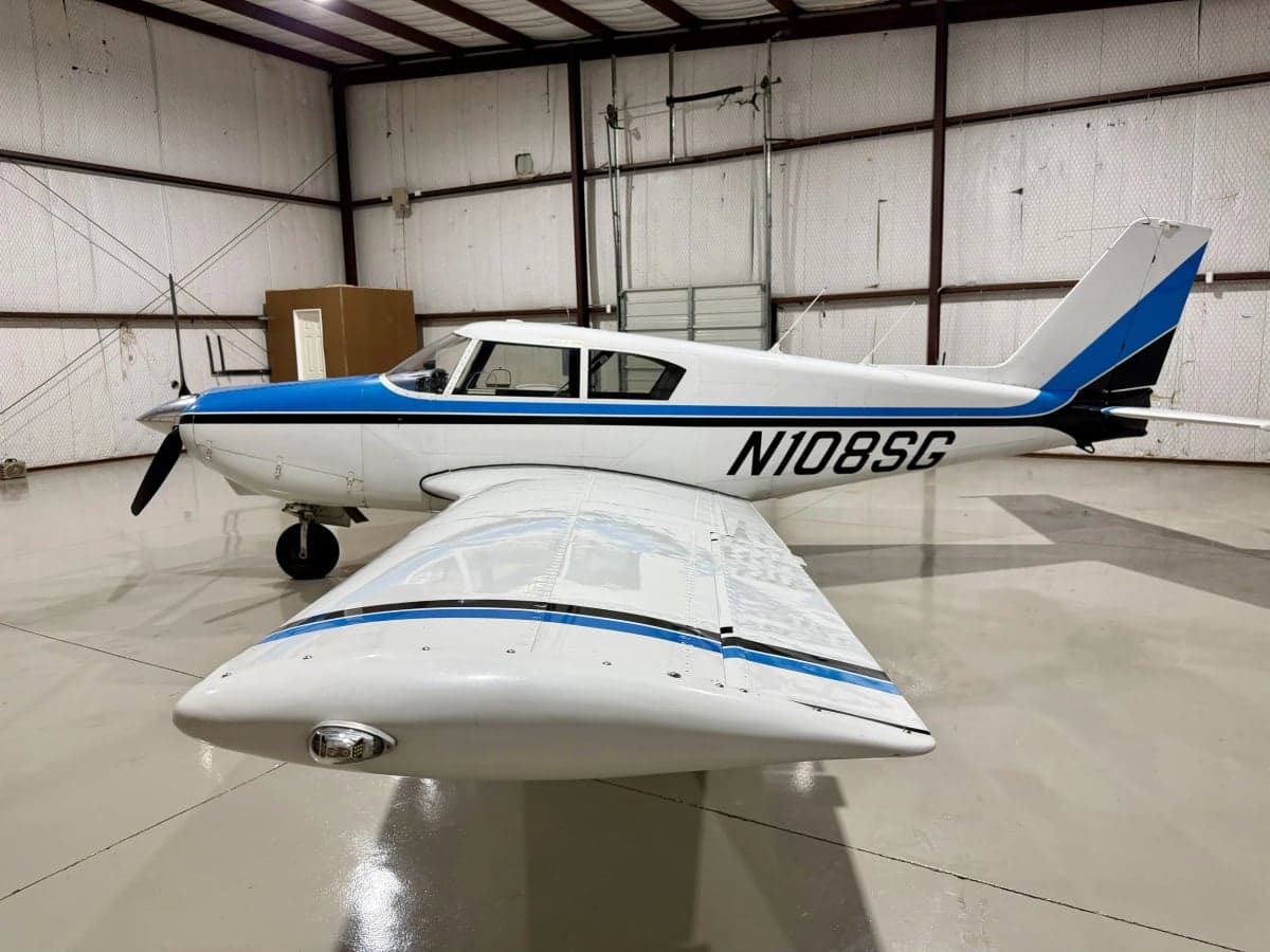 A white and blue Piper PA-28-140 Cherokee is parked in a hangar, showing the nose and wing.