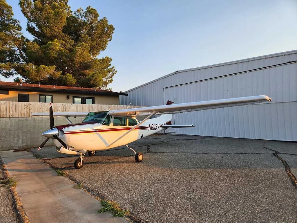 A white and maroon Cessna 172 aircraft, N6113V, is parked on an asphalt apron next to a hangar at sunset.