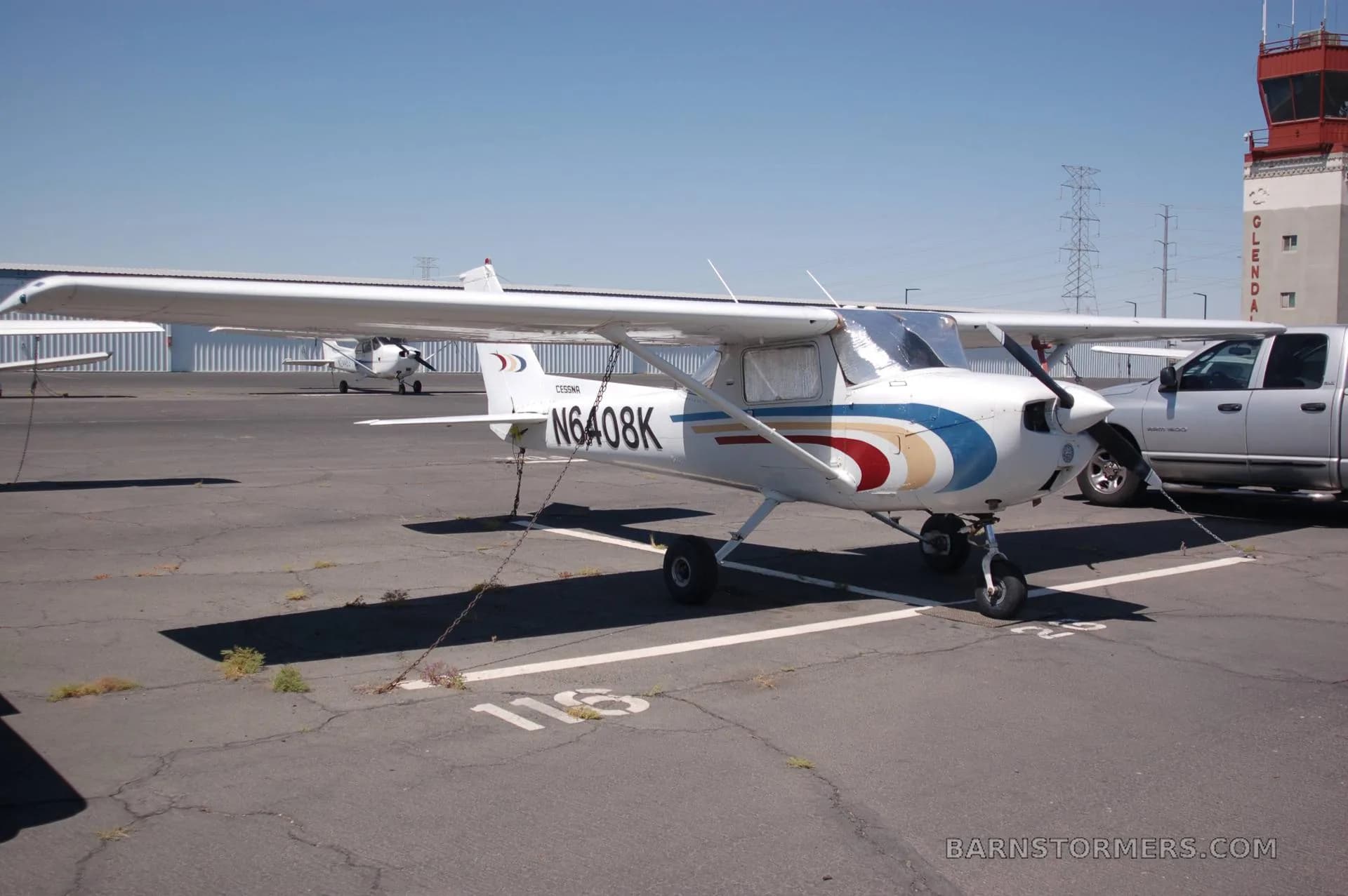 A white Cessna N6408K with blue, gold, and red stripes is tied down on an airfield next to a control tower.