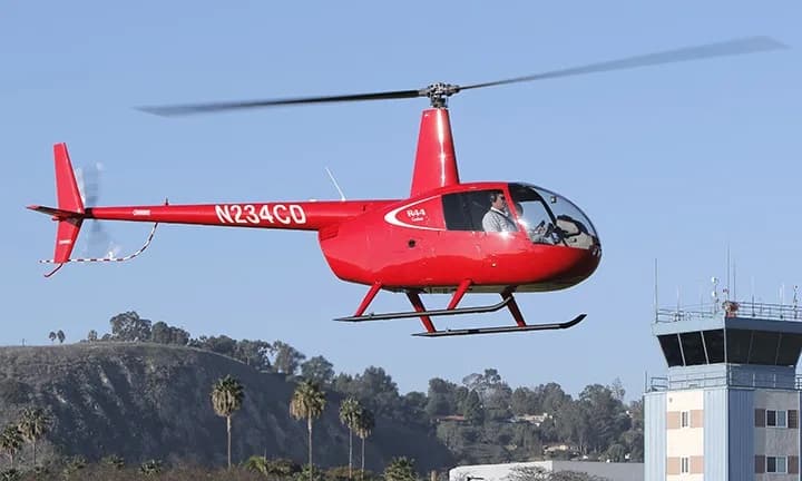 A red Robinson R44 helicopter, registration N234CD, flies near an airport control tower.