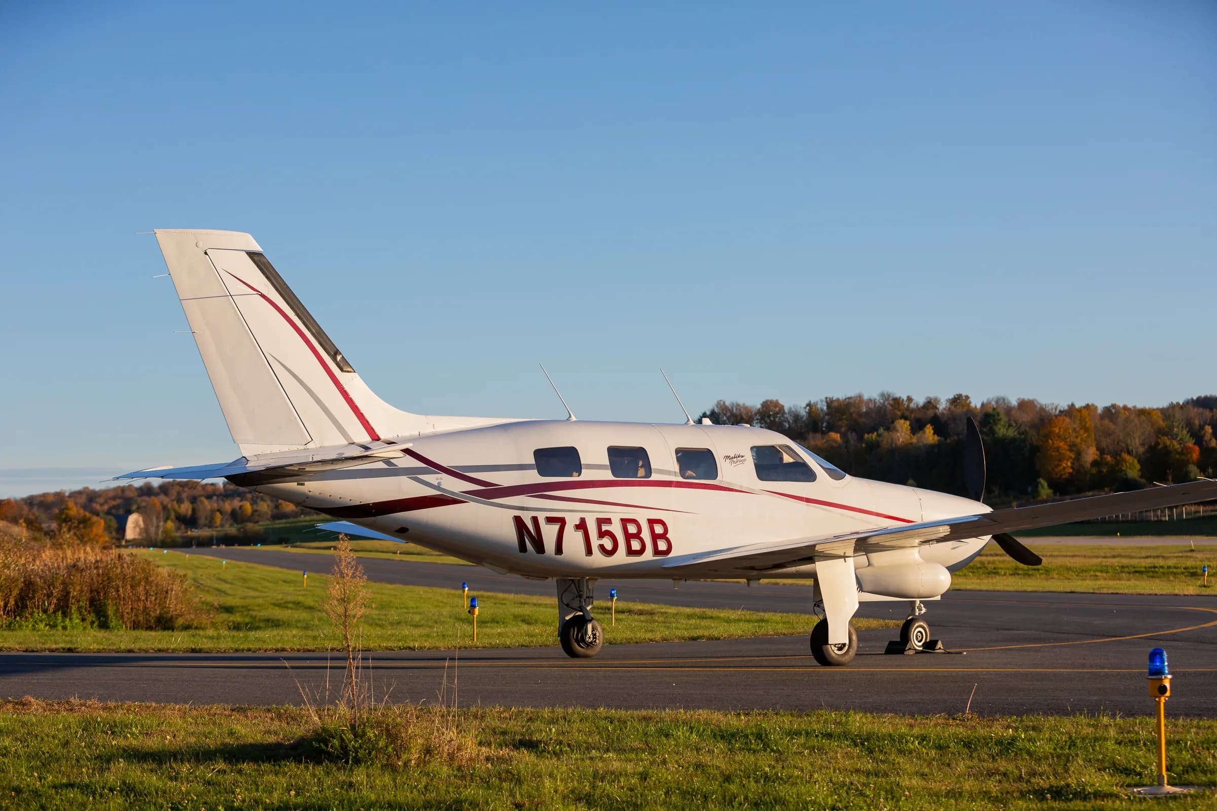 A side view of the Piper Malibu Mirage N715BB parked on a runway with autumn trees in the background.