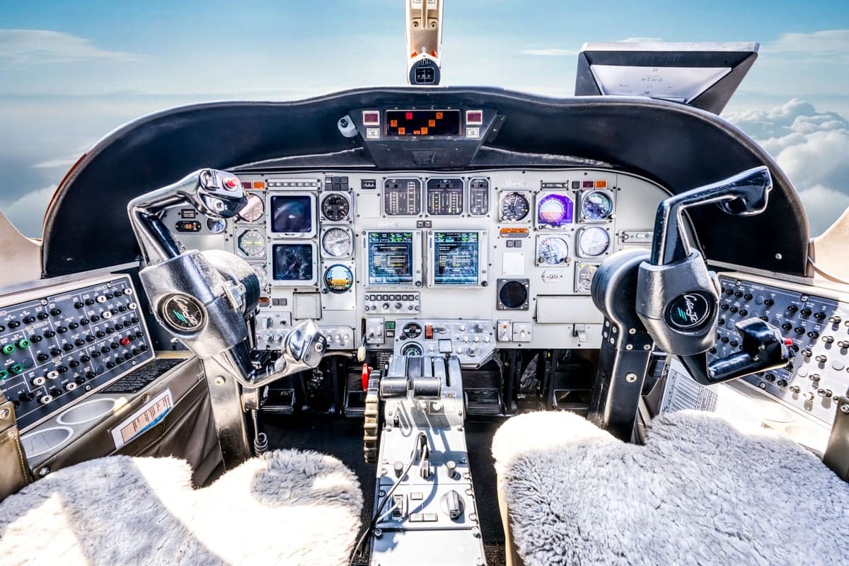 The cockpit of a jet aircraft features a complex instrument panel with multiple screens and controls, and two sheepskin-covered pilot seats.