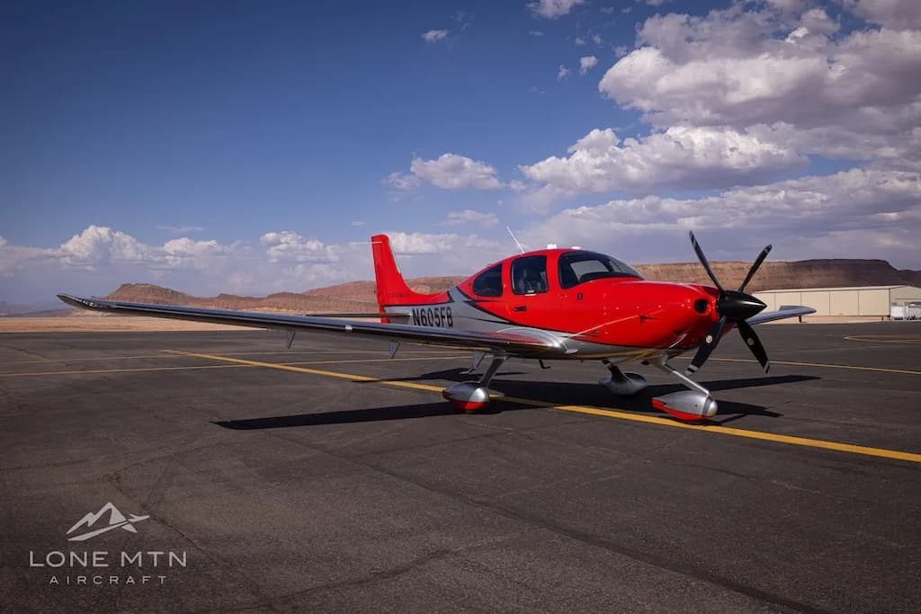 A front-quarter view of a red and silver Cirrus SR22T aircraft (N605FB) on an airfield with clouds in the sky.