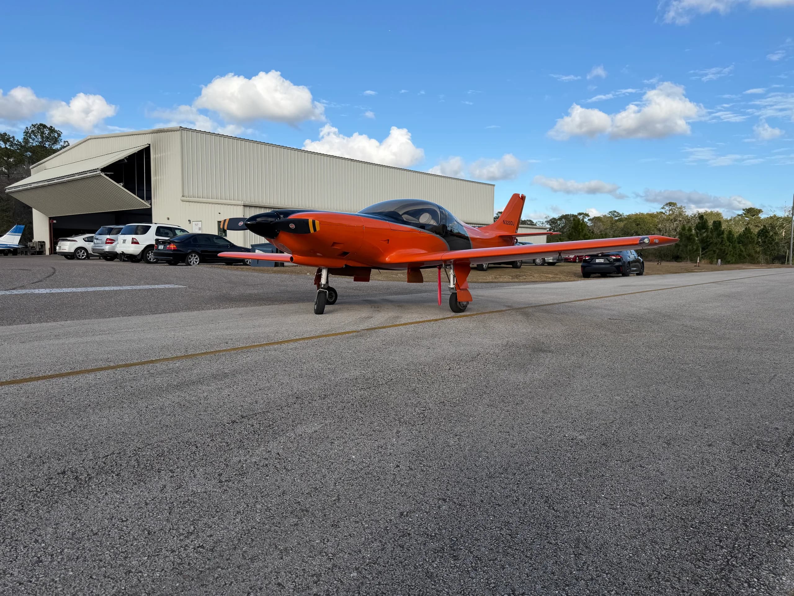 An orange and black experimental aircraft, N320DJ, sits on an airfield tarmac in front of a hangar under a partly cloudy sky.