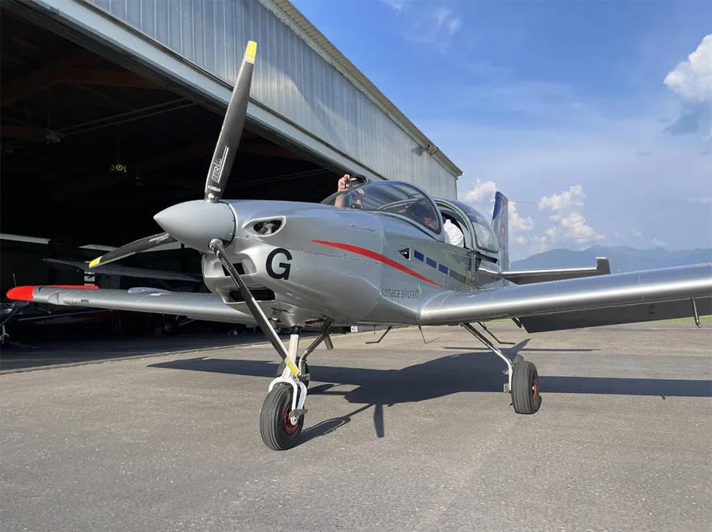 A silver Sonaca 200 aircraft with red accents is parked in front of an open hangar.