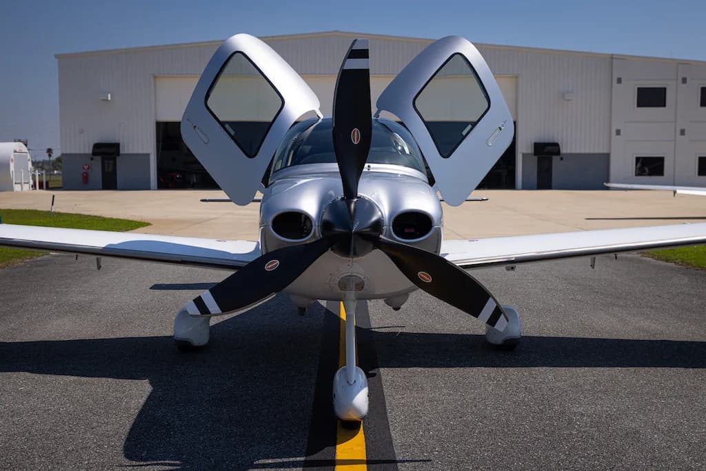 A head-on view of the silver Cirrus SR22 aircraft N904BL with its doors open, parked in front of a hangar.