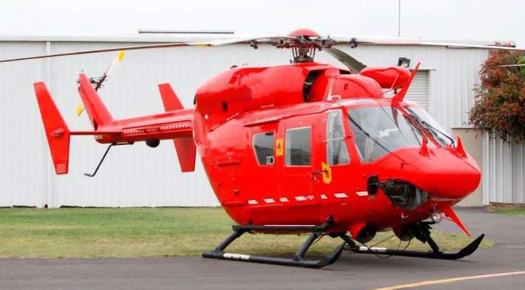 A red helicopter with skids parked on an asphalt surface next to a white hangar building.