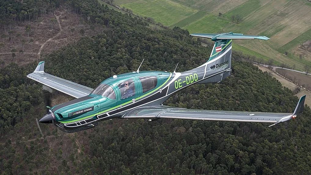 A green and black Diamond DA40 NG aircraft with registration OE-DDD flying over a forested landscape.