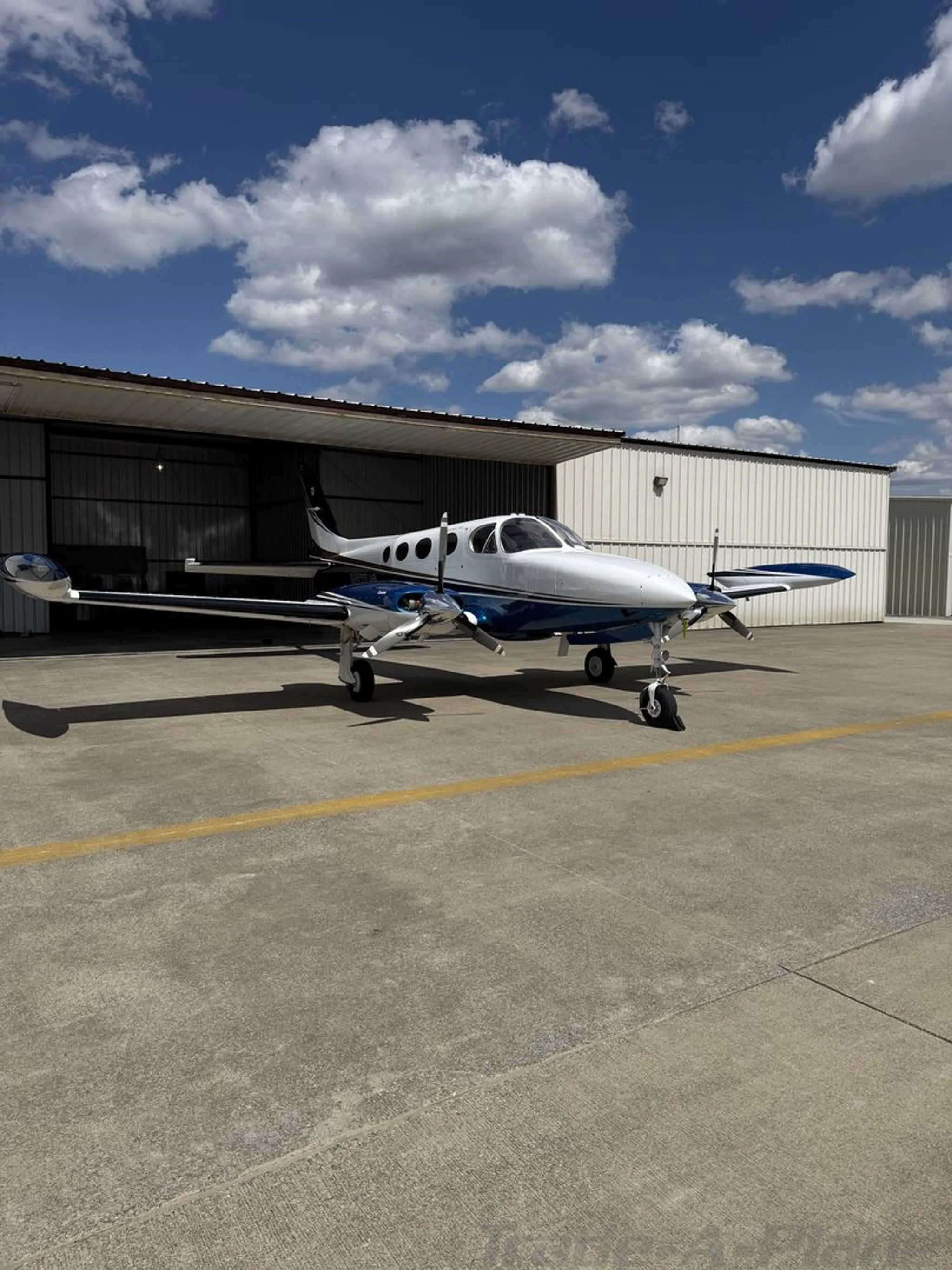 A white, blue, and gold Cessna 414 twin-engine aircraft parked on an apron in front of a hangar.