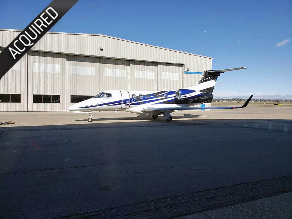 A white and blue business jet is parked on an airport tarmac in front of a hangar under a clear sky.