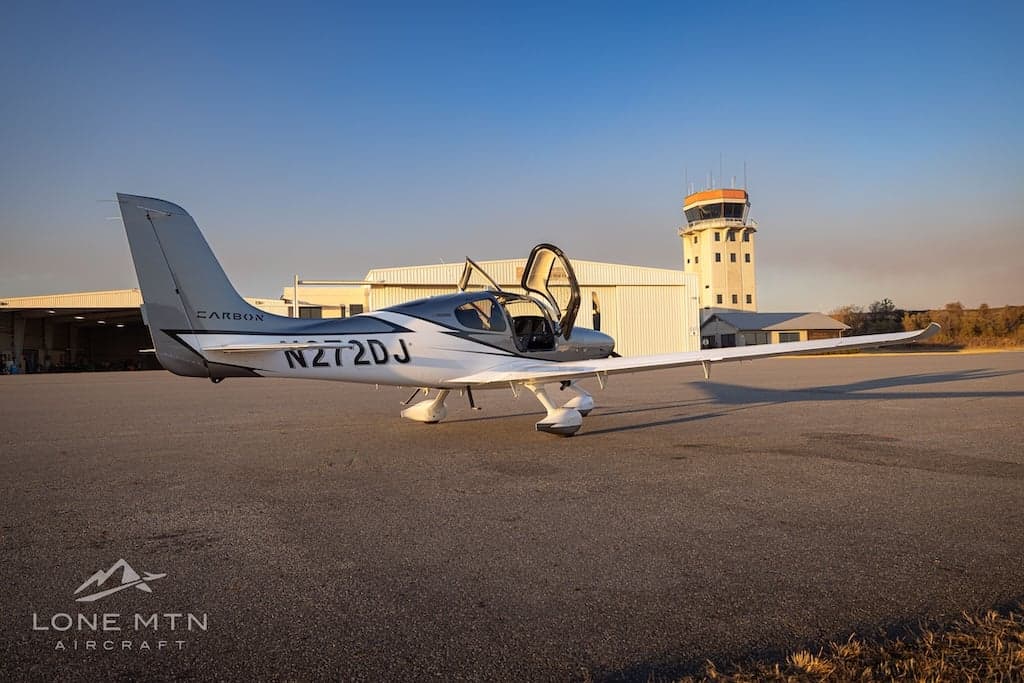A Cirrus SR22T aircraft with open doors is parked on the tarmac near an airport control tower.