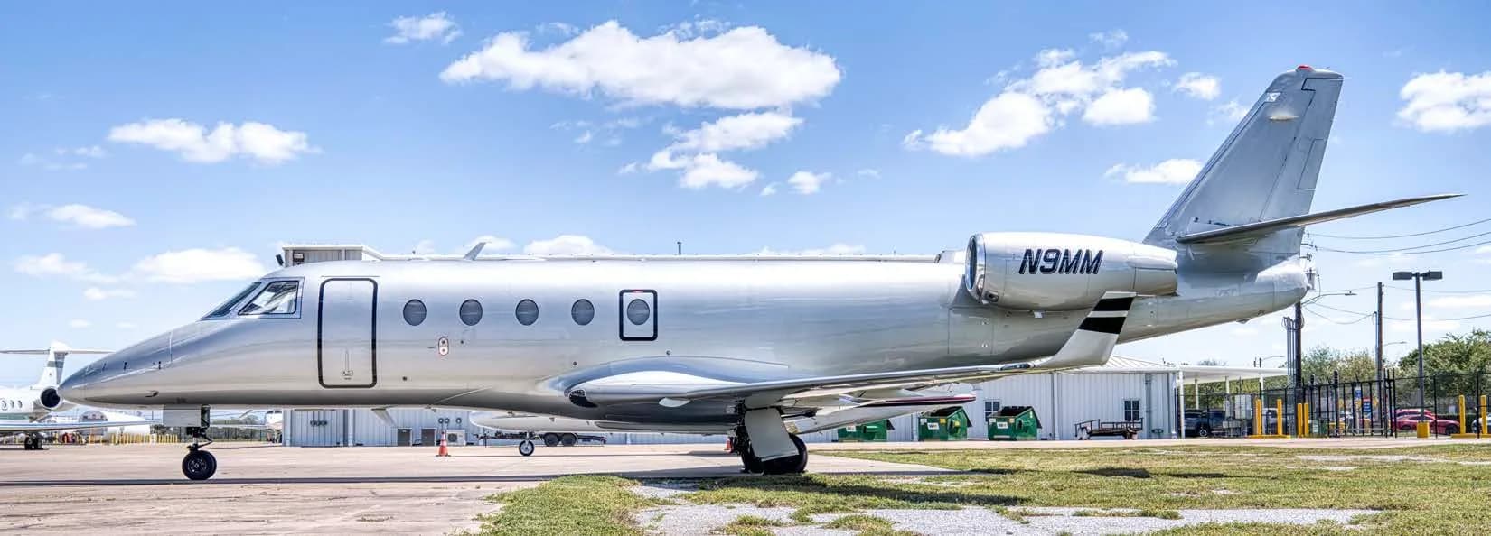 A silver business jet with registration N9MM parked on an airfield under a blue sky.