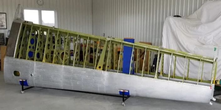 An aircraft wing section, with its internal ribs and spars exposed on one side, resting on supports in a hangar.