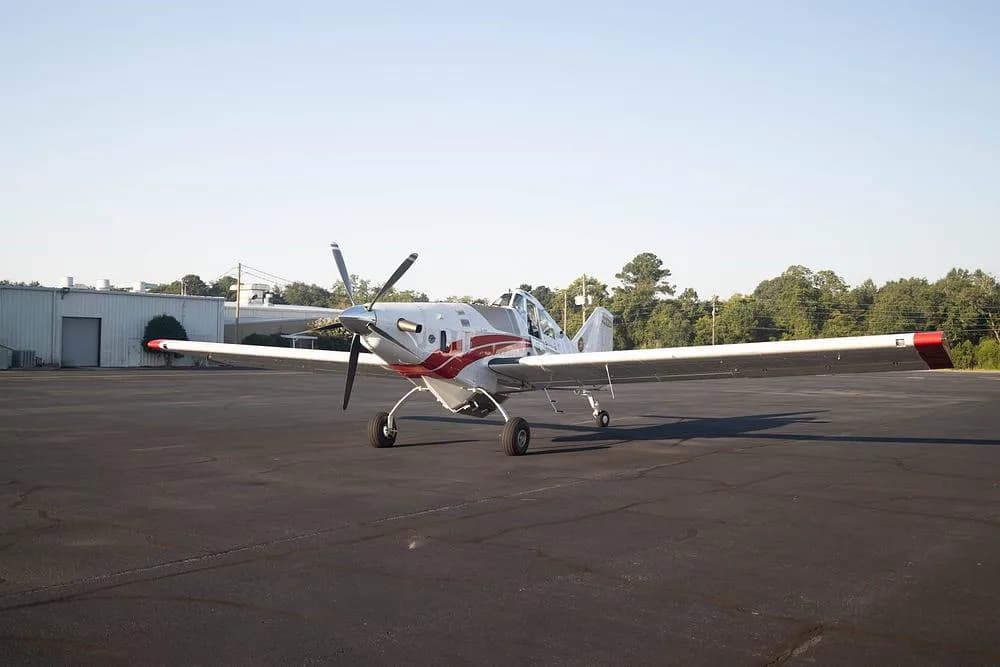 A white and red Thrush 510G aircraft parked on an asphalt tarmac with trees and buildings in the background.