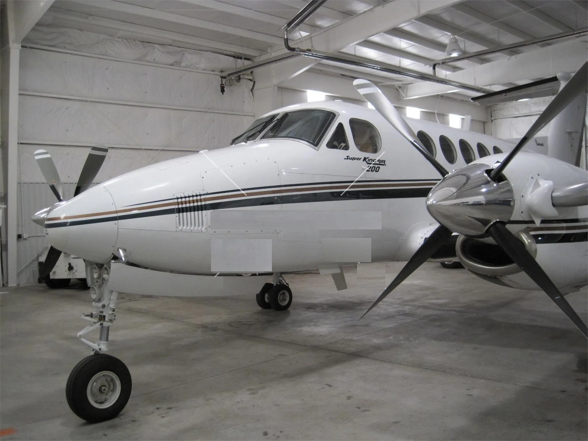 A white Beechcraft Super King Air 200 twin-turboprop aircraft with brown and black stripes, parked inside a hangar.