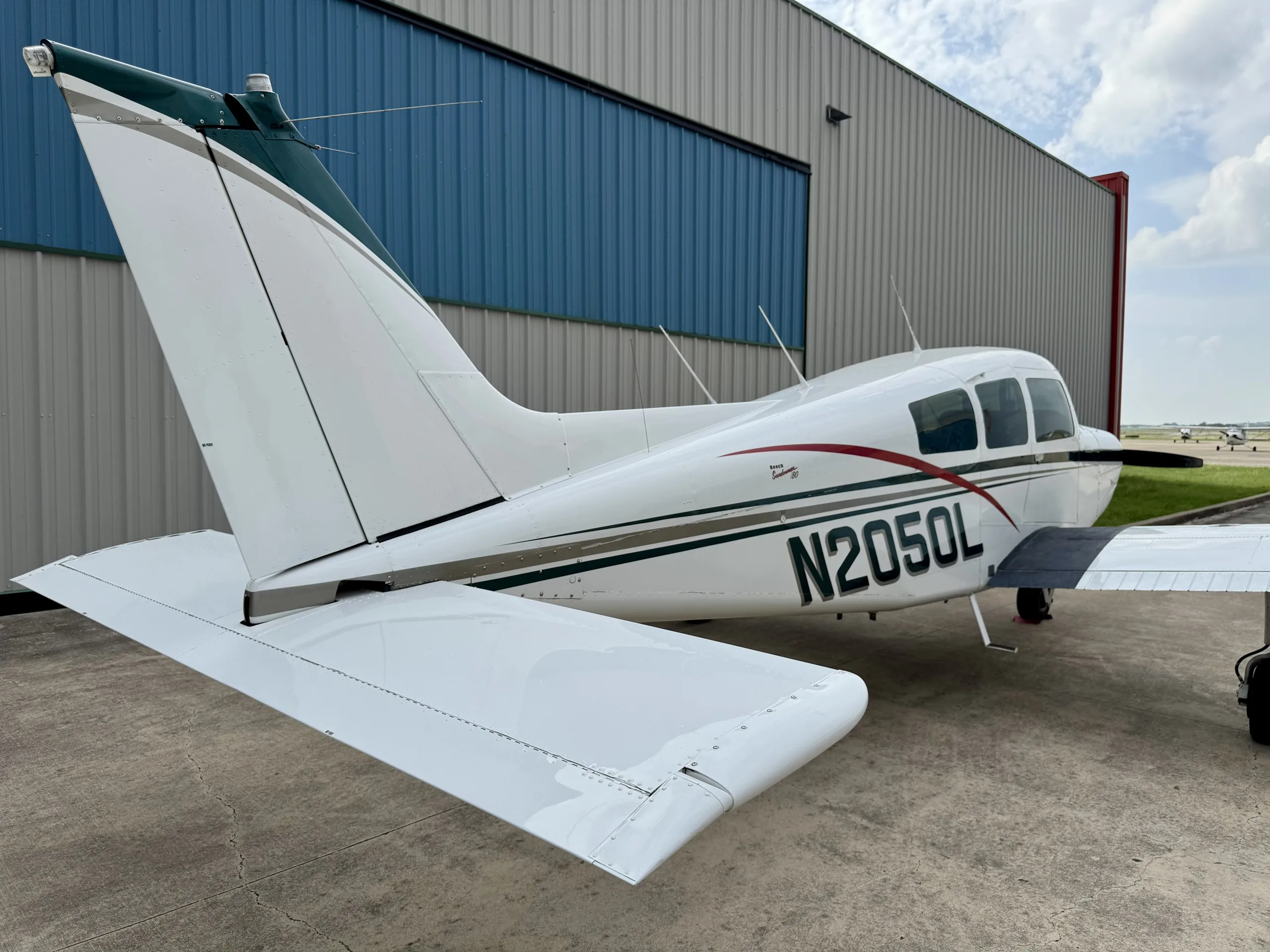 Rear three-quarter view of a white and green Beechcraft Sundowner N2050L with other aircraft in the background.