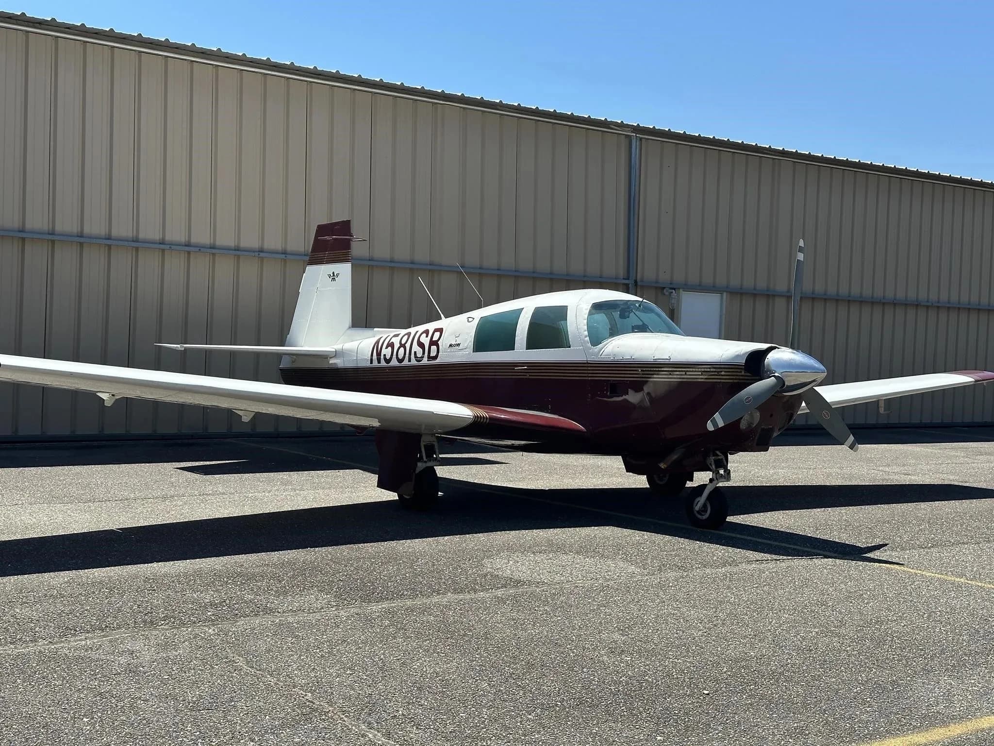 A white and maroon Mooney M20 Mark 21 aircraft, registration N581SB, parked on an asphalt ramp next to a hangar.
