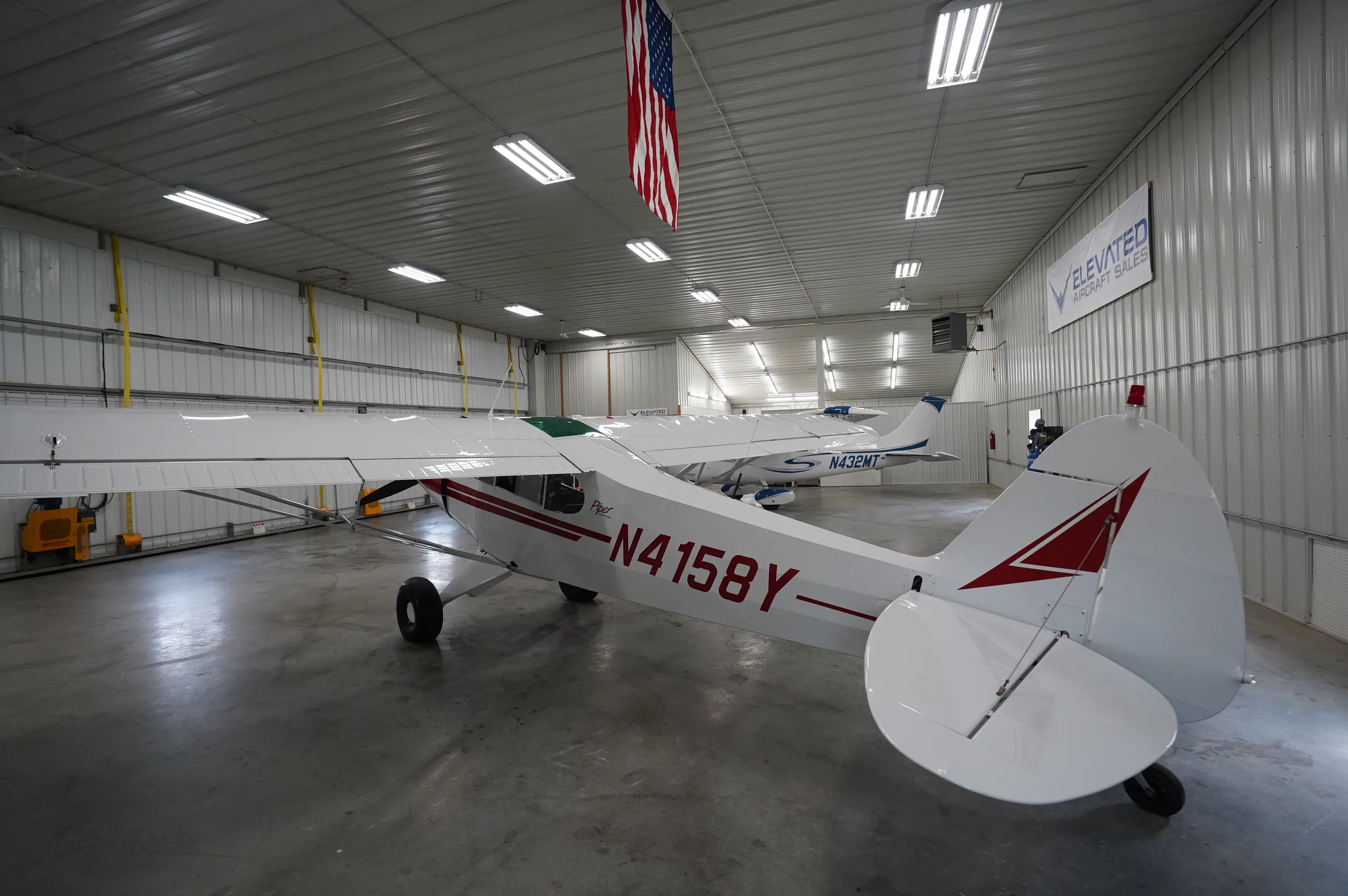 The rear view of a white Piper Cub aircraft, registration N4158Y, with red tail markings, parked in a hangar with another aircraft in the background.