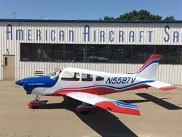 A white, blue, and red single-engine aircraft N5587V is parked on a tarmac in front of the American Aircraft Sales building.