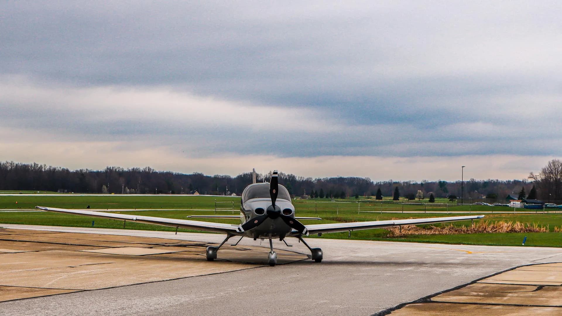 A silver and white Cirrus SR22 aircraft is seen from the front, parked on an airport tarmac under a cloudy sky.