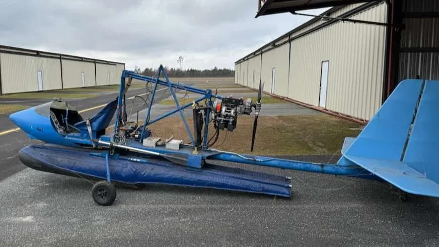 A blue ultralight aircraft on floats, with its tail and engine visible, parked on an airfield next to a hangar.