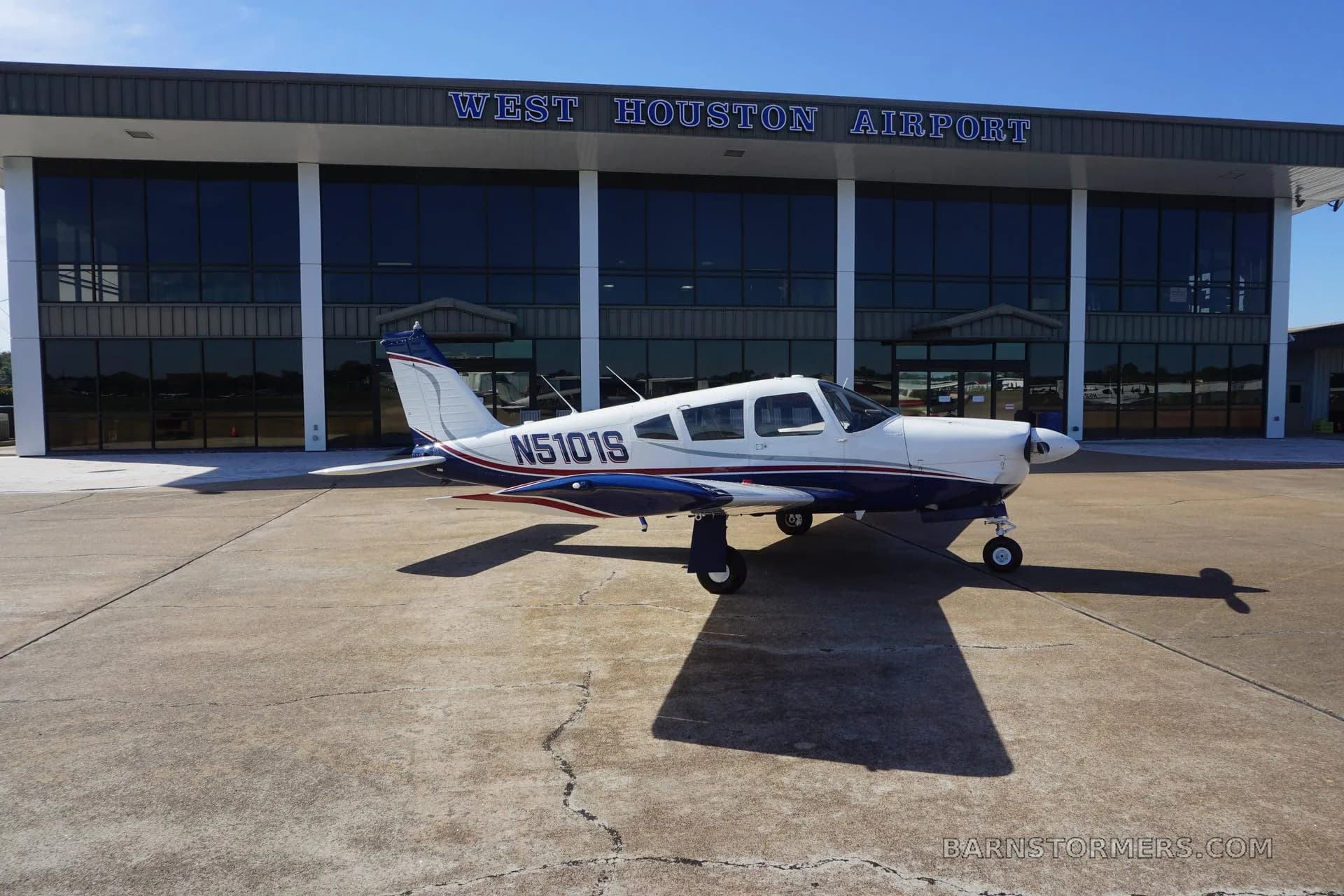 A white and blue Piper Cherokee aircraft with red accents is parked on the tarmac in front of the West Houston Airport terminal building.