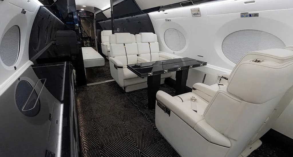 A view inside the business jet cabin showing white leather seats, a black table, and dark patterned carpet.