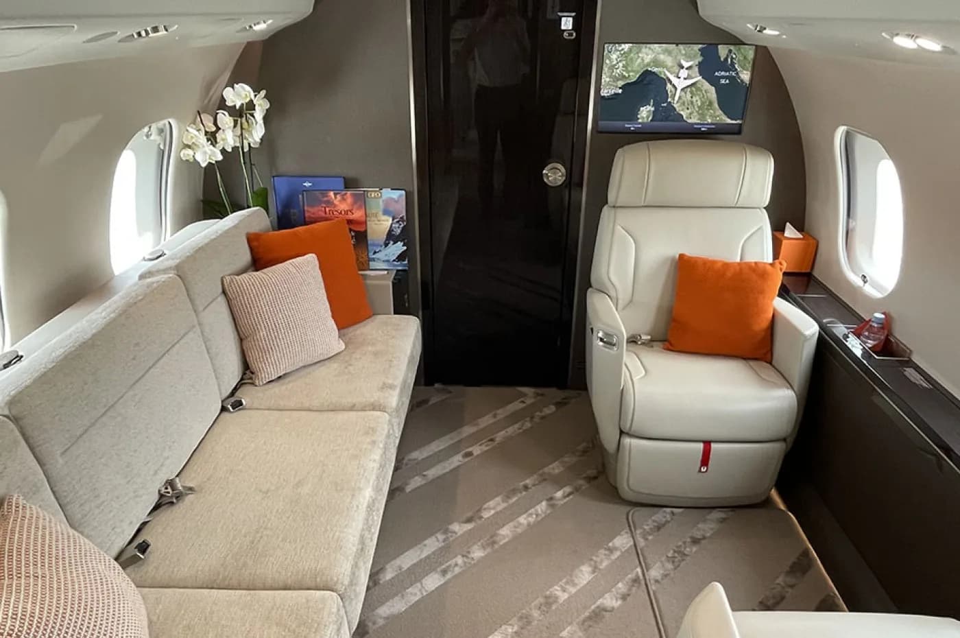 The spacious interior of a private jet, featuring a long sofa with orange pillows, a single armchair, and a wall-mounted screen displaying a map.