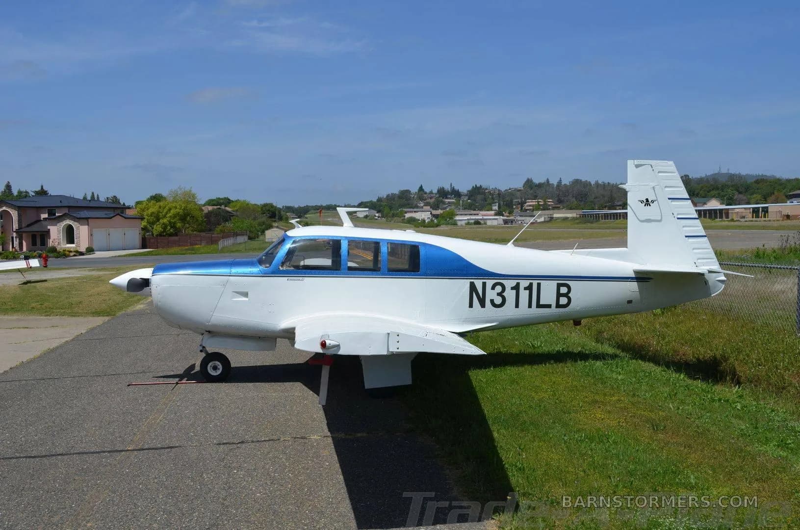A white and blue single-engine propeller aircraft, N311LB, is parked on an asphalt ramp next to grass under a clear sky.