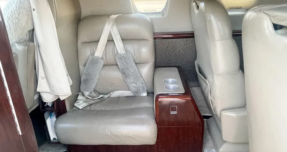 A tan leather passenger seat with sheepskin shoulder pads and a wooden side console with cup holders in the aircraft cabin.