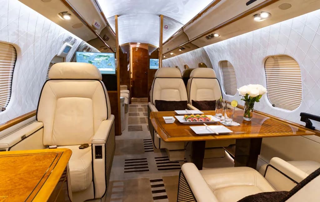 A wide view of the private jet's luxurious cabin with beige seats, a wooden table with sushi, and a map on a screen.