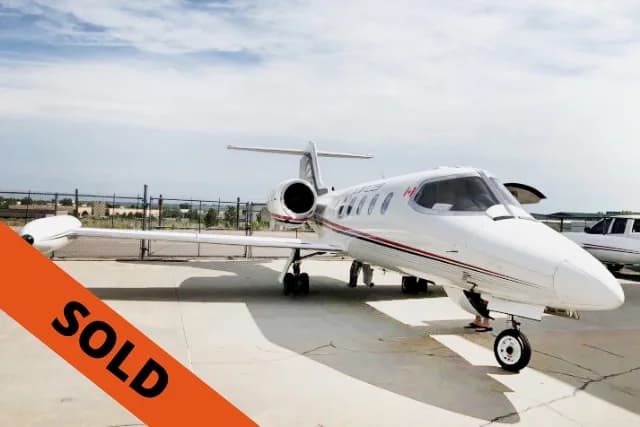 A white business jet with red and black stripes and a Canadian flag on the nose, parked on an airfield with its door open.
