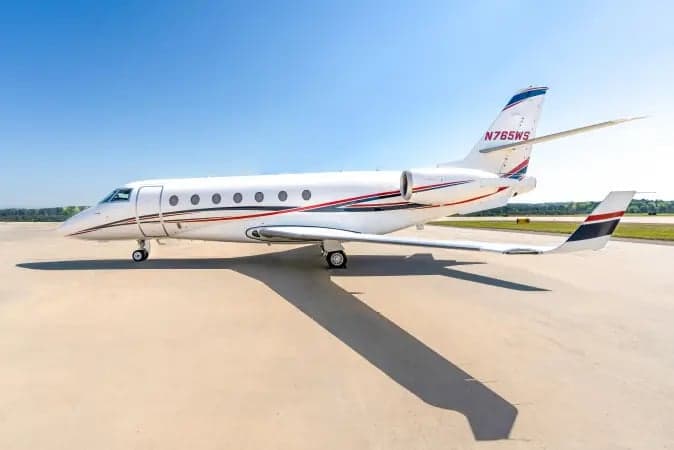 A white business jet with red and blue stripes, registration N765WS, parked on an airfield under a clear blue sky.