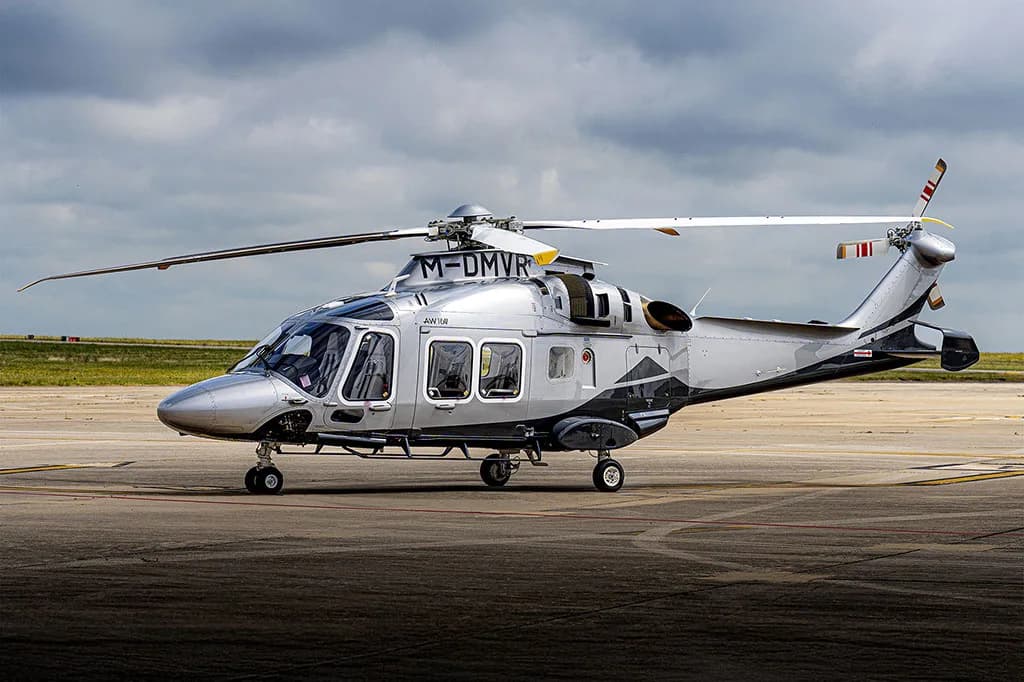 A silver and dark grey AgustaWestland AW169 helicopter (M-DMVR) parked on an airfield under a cloudy sky.