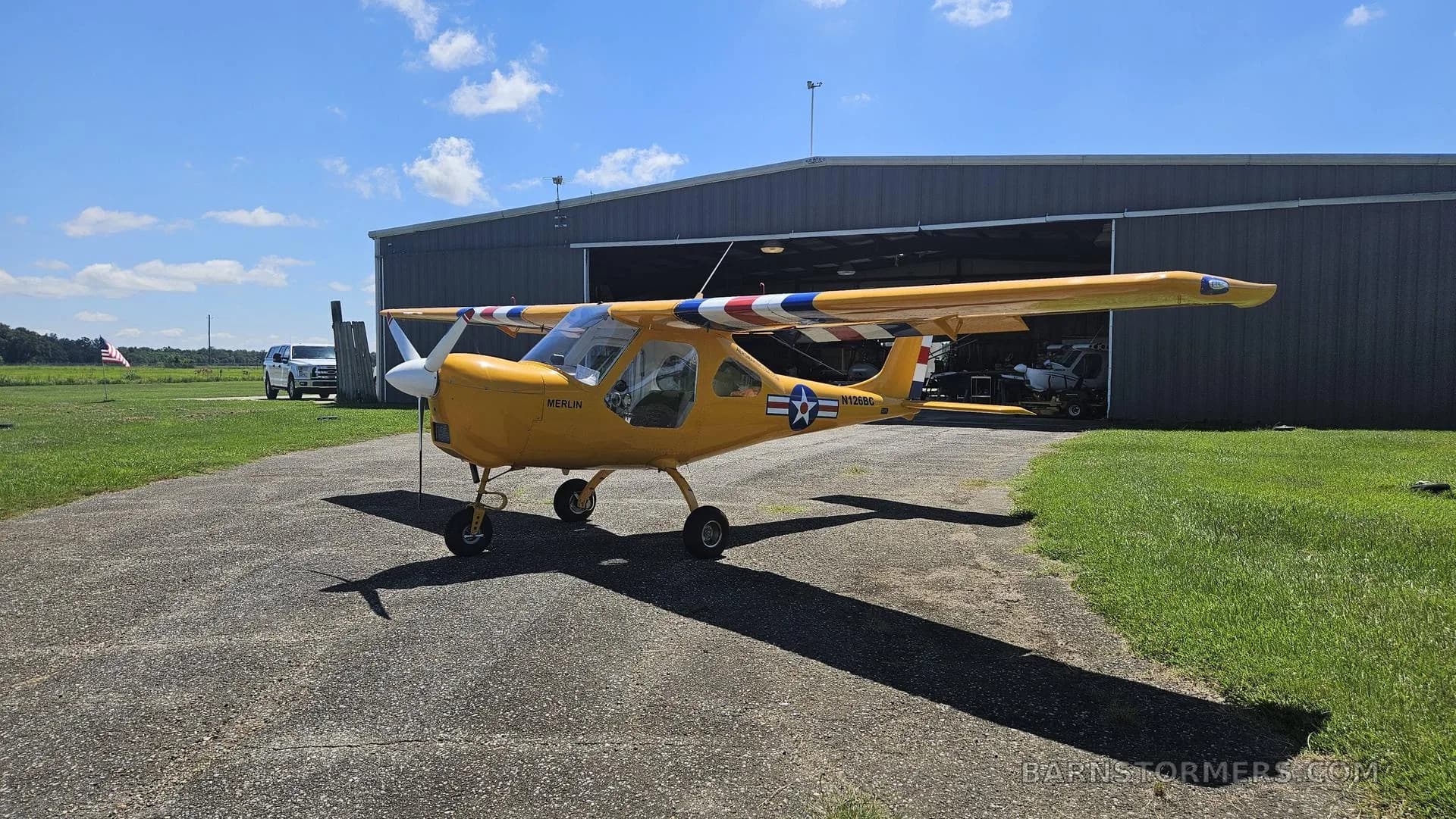 A yellow Merlin ultralight aircraft with a red, white, and blue striped wing is parked on a concrete pad outside a gray hangar on a sunny day.