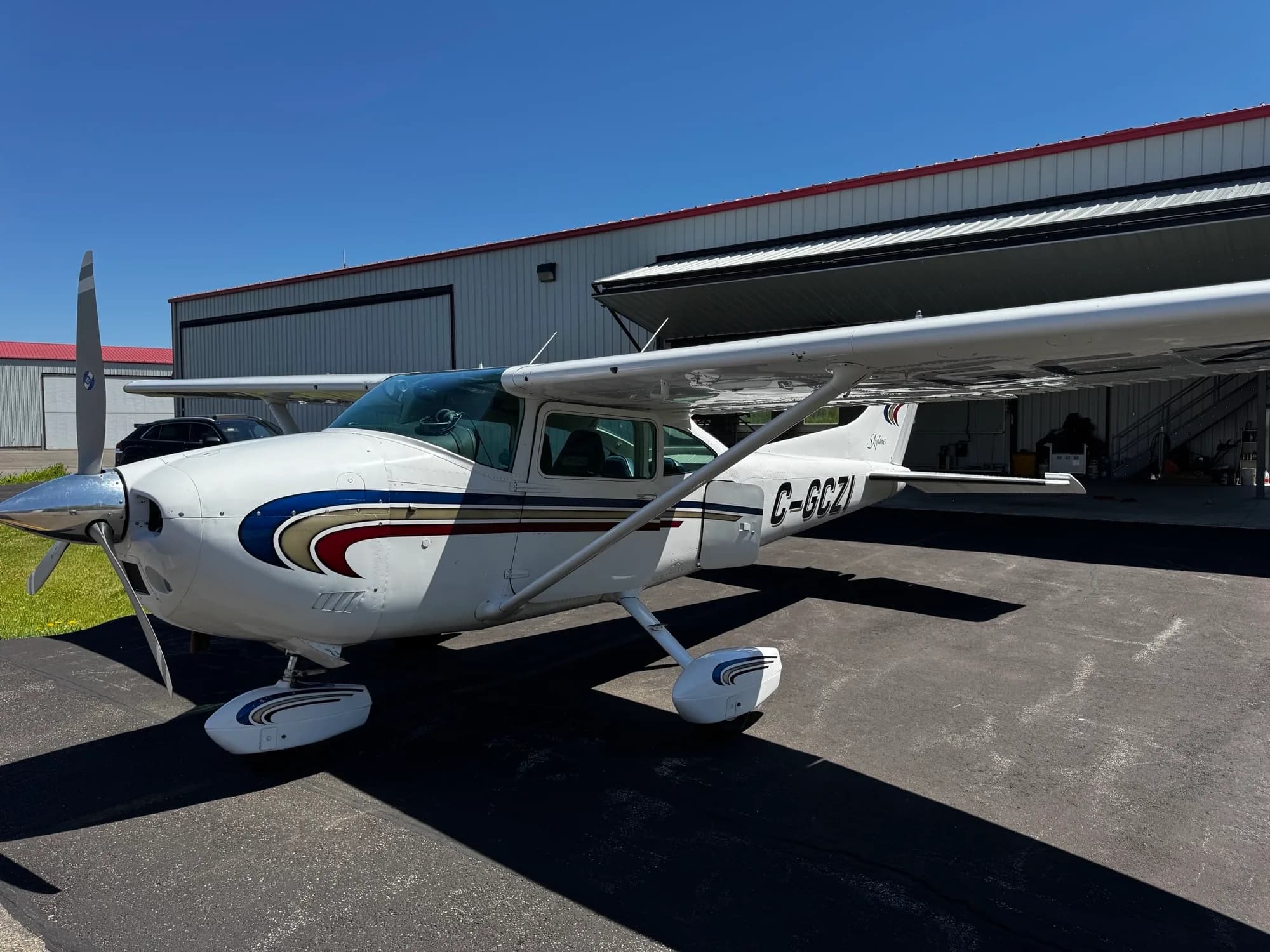 A white Cessna Skylane C-GCZI with blue, gold, and red stripes, parked on an asphalt ramp under a clear blue sky.