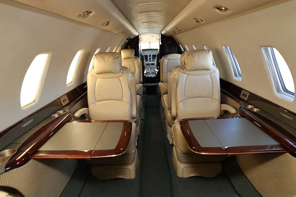 The interior cabin of a private jet showing beige leather seats with pull-out tables extended from the dark wood side panels.
