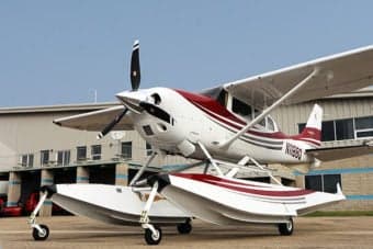 A white and blue Cessna 206 on floats is docked on a lake with mountains in the background.
