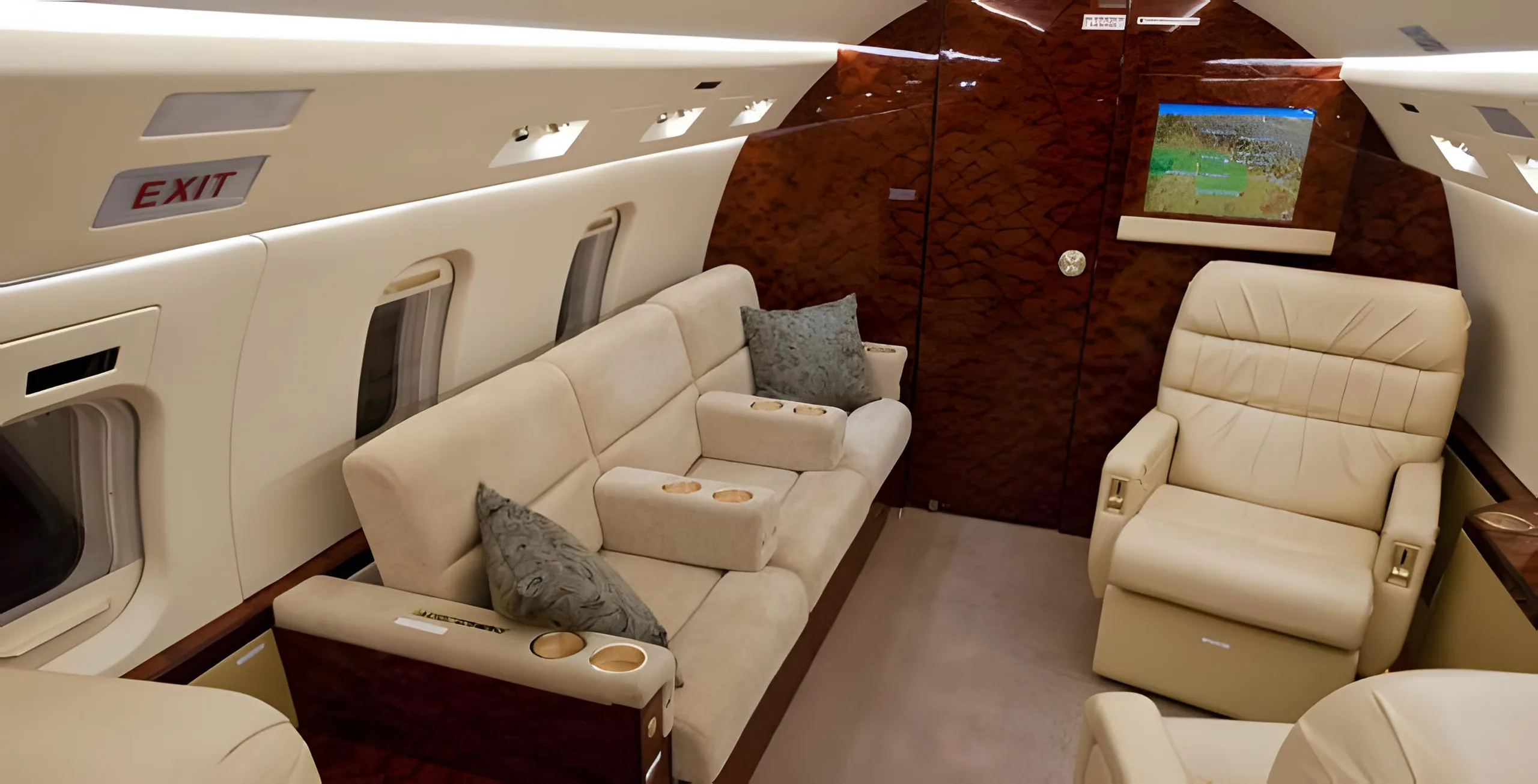 A view of the aircraft's cabin interior featuring a long beige sofa with pillows and cup holders, a single beige leather seat, and a map on a screen.