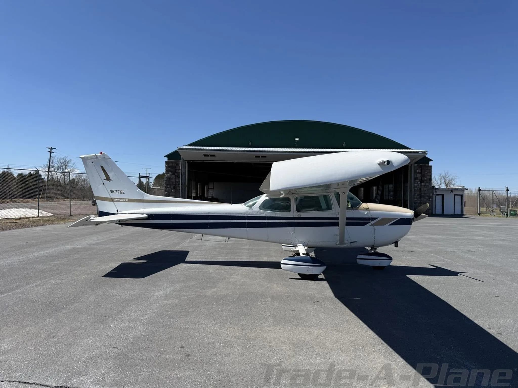A side view of the Cessna 172 aircraft N6778E, white with blue and tan stripes, parked on an airfield.