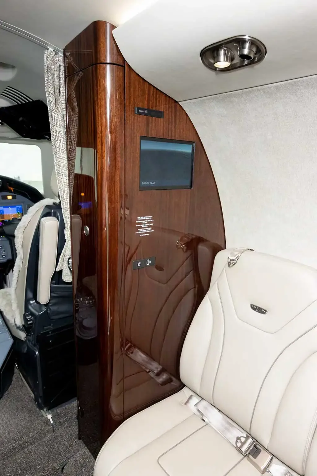 A dark wood-paneled cabinet with a small screen is positioned next to a light tan leather seat in the private jet cabin.