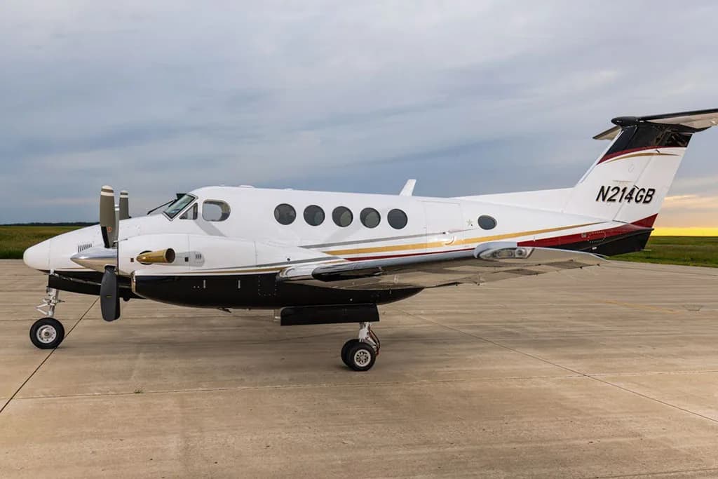 A white Beechcraft King Air N214GB with black, gold, and red stripes parked on an airfield under a cloudy sky.
