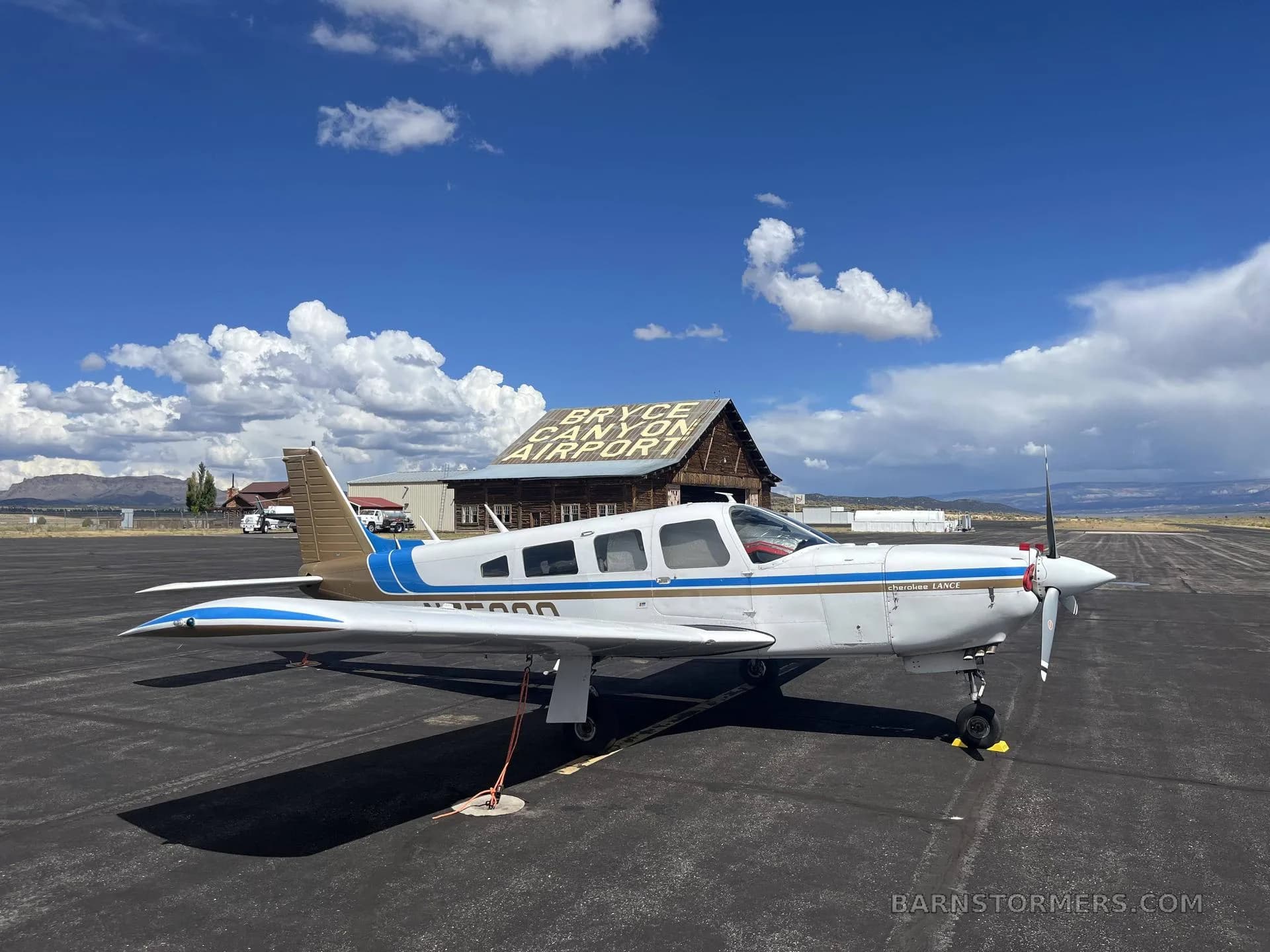 A white and blue Piper Cherokee Lance (N75398) parked on an asphalt ramp in front of the "Bryce Canyon Airport" building under a partly cloudy sky.