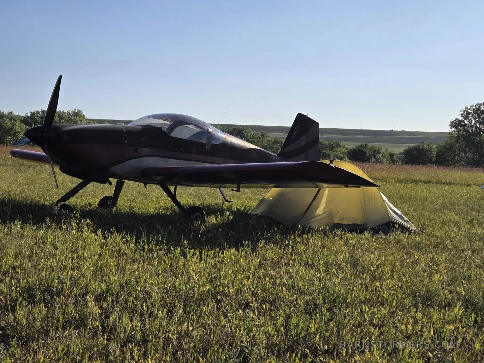 A dark-colored N4361V aircraft with a red stripe on the tail, parked in a grassy field next to a yellow tent.