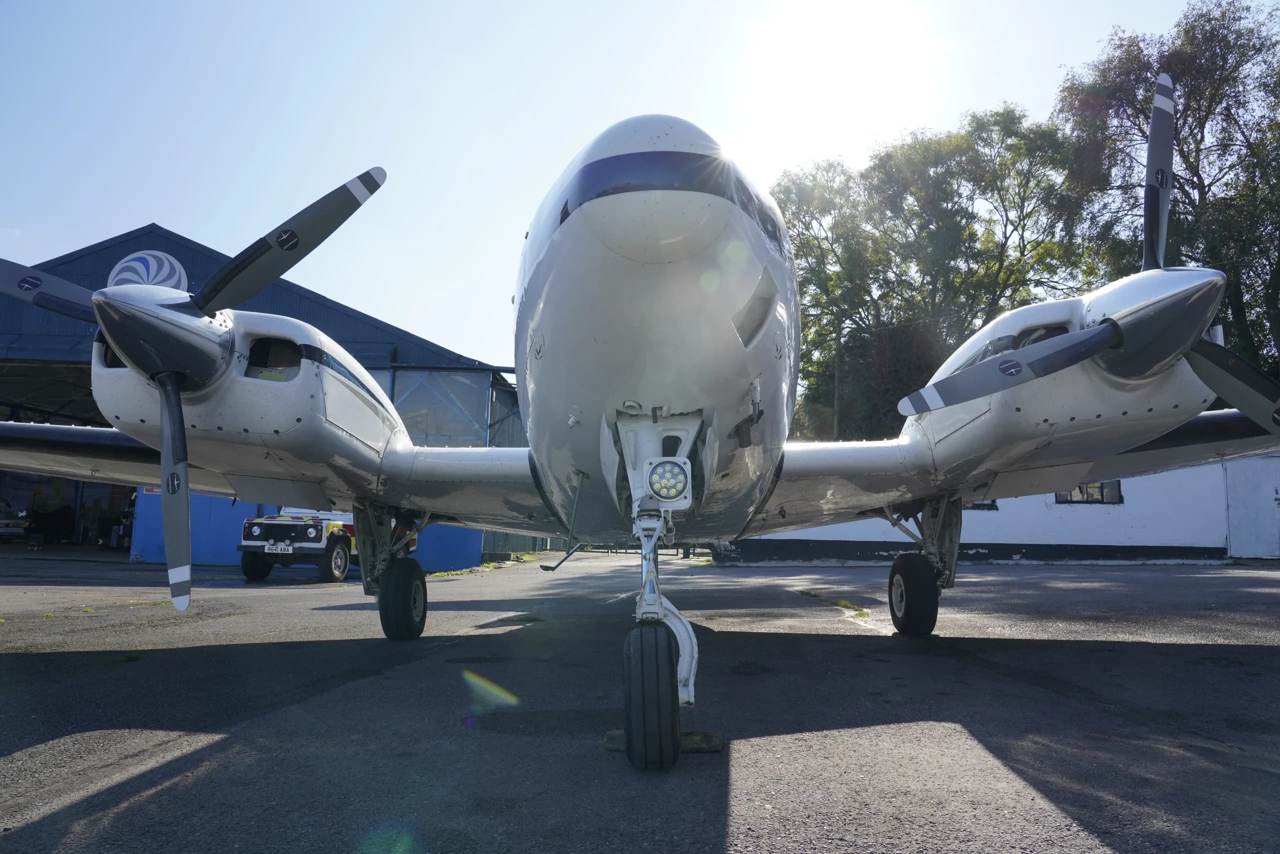 A head-on view of a white twin-engine propeller aircraft with blue stripes, parked on an asphalt apron.
