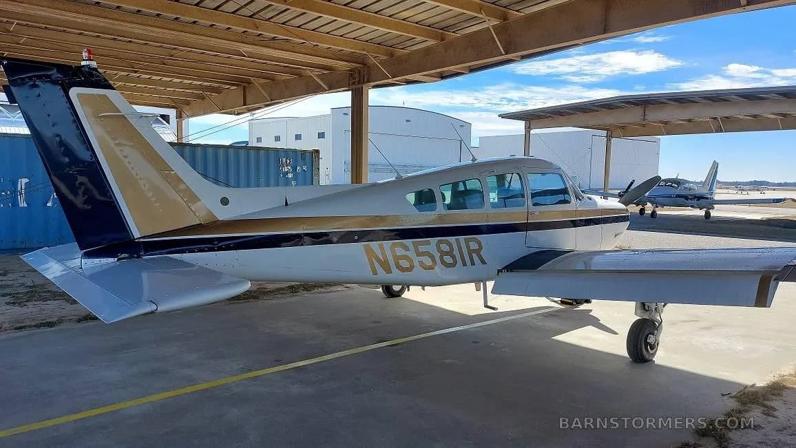 A light-colored single-engine aircraft with black and gold stripes, tail number N6581R, parked under a hangar.