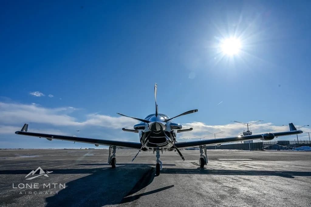 A low-angle front view of a TBM 940 aircraft parked on an asphalt ramp, with the sun brightly shining overhead.