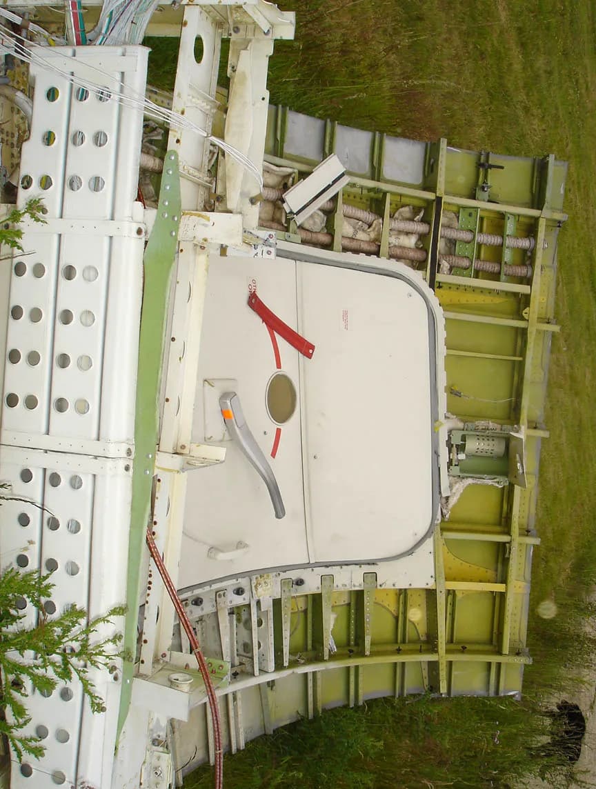 A side view of a disassembled aircraft fuselage section with an emergency exit door and exposed framework.
