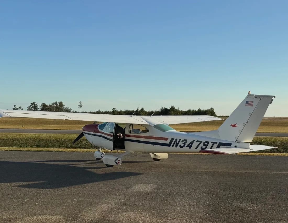 A white Cessna 182 with red and blue stripes, registration N3479T, parked on an asphalt runway with its door open.