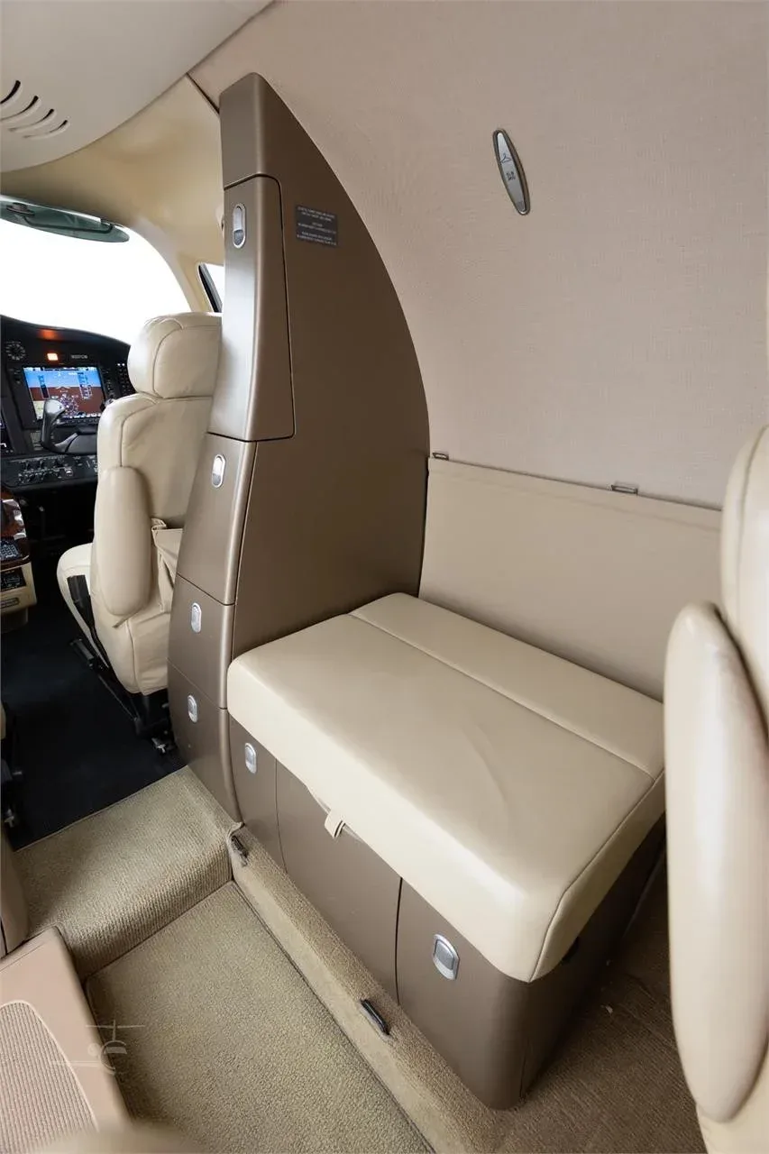 A view inside the Cessna Citation M2 cabin showing a beige bench seat and storage compartments, with the cockpit visible in the background.